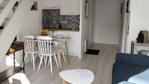 Uma cozinha compacta com armários brancos e painel de fundo estampado, uma área de refeições com cadeiras brancas e uma mesa pequena, e uma escada que leva a outro nível.