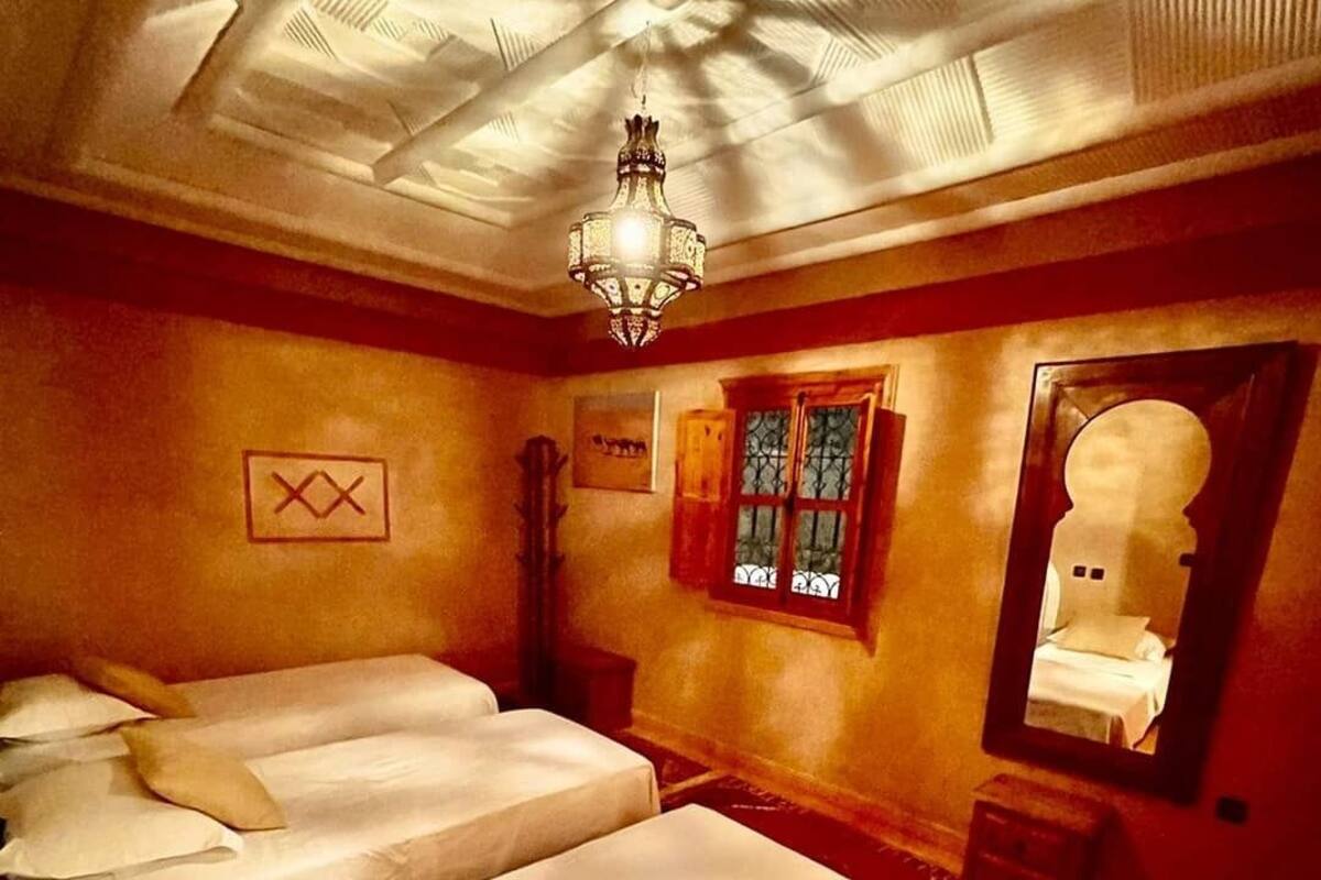 A room with three beds, a chandelier, a window with iron bars, and a mirror reflecting a bed.