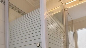 Casa de banho com chuveiro em cabine de vidro, lavatório branco e espelho.