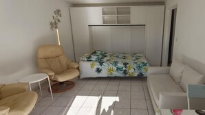 Um espaço compacto com sofá, poltrona, mesa pequena, cama com colcha estampada com tema tropical e guarda-roupa com vários compartimentos.