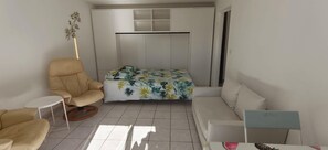 A compact living space with a sofa, a chair, a small table, a bed with a tropical print bedspread, and a wardrobe with multiple compartments. - Quiet apartment with balcony and swimming pool (Fréjus)