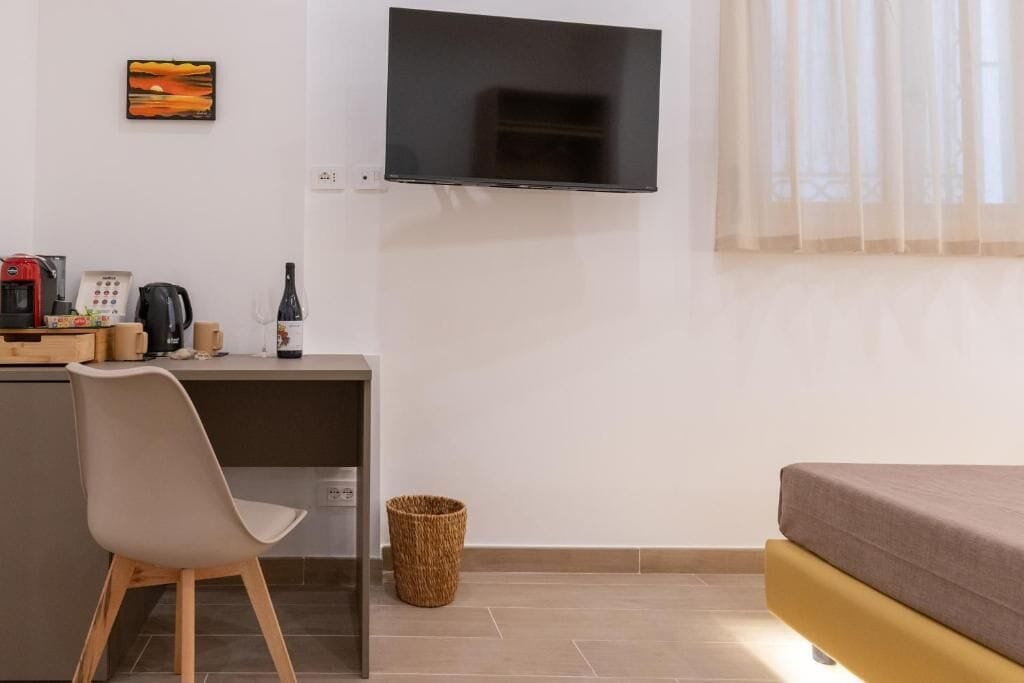A room with a bed, a desk with a chair, a television, a coffee machine, a wine bottle, and a basket.