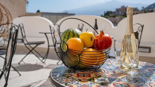 A table with a fruit bowl, a bottle of champagne, and glasses on a patterned tablecloth. - Light View (Taormina)