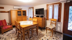 Een woonkamer met een houten kast, een televisie op een standaard, een eettafel met stoelen, een bank en een vloer met patroon.