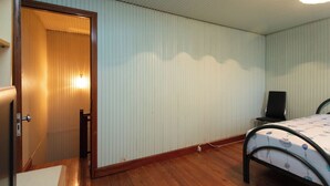 Een kamer met een houten deur, een bed met een beddengoed met een patroon, een stoel en een wandlamp.