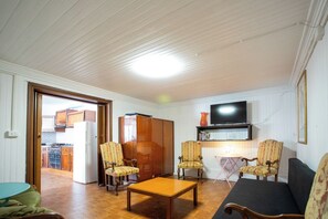 A living room with a sofa, two armchairs, a wooden coffee table, a television mounted on the wall, and a kitchen visible through an open door.