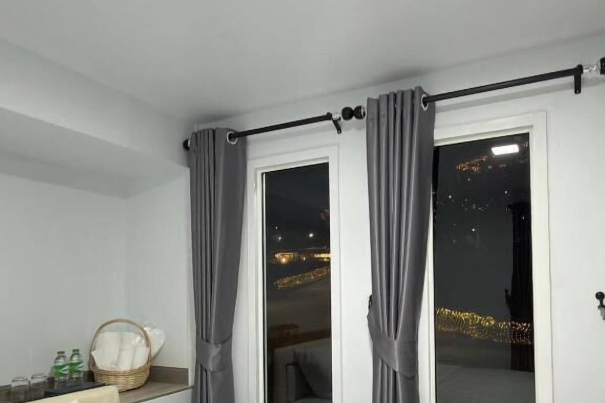 A hotel room with a bed, a window with curtains, a night view outside, and a small table with a basket and bottles.
