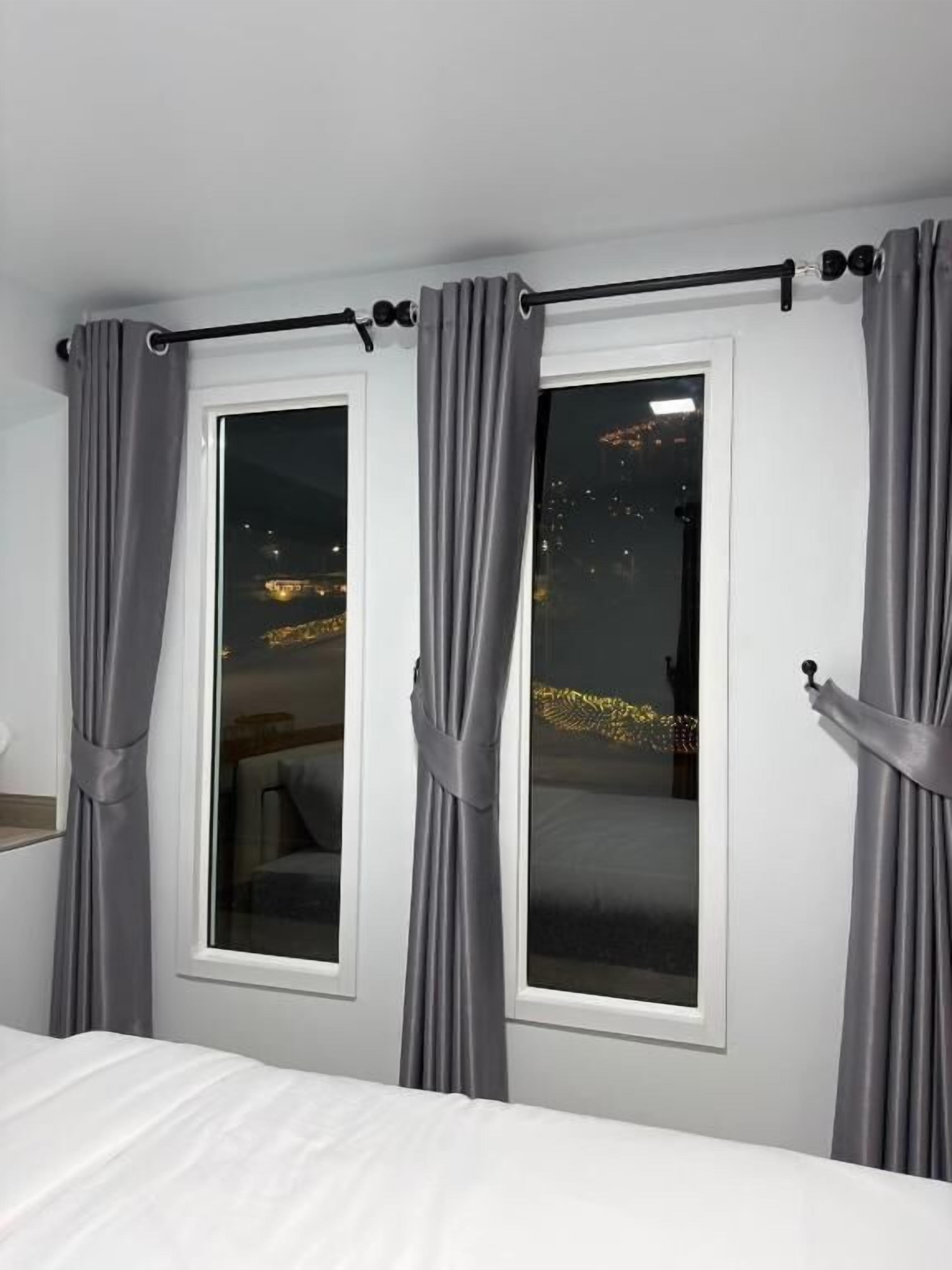 A bedroom with two windows, grey curtains, a bed, and a view of a cityscape at night.