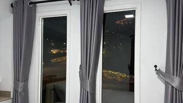 A bedroom with two windows, grey curtains, a bed, and a view of a cityscape at night.