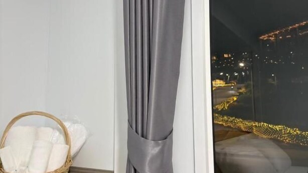 A room with a grey curtain, a wicker basket with white linens, and a view of a cityscape at night. - Siriwan boutique hotel (Takua Thung)