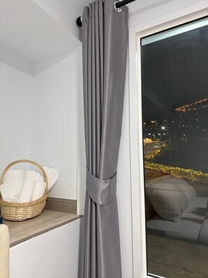 A room with a grey curtain, a wicker basket with white linens, and a view of a cityscape at night. - Siriwan boutique hotel (Takua Thung)