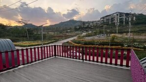 A wooden deck with a red and pink railing, overlooking a hilly landscape with buildings and a road. - Siriwan boutique hotel (Takua Thung)