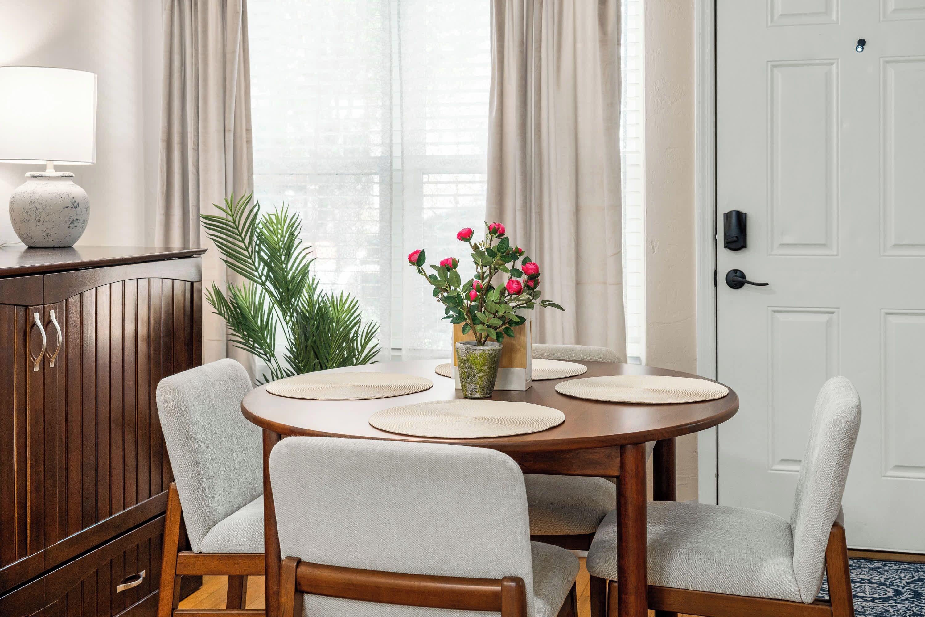 Un comedor con una mesa redonda, cuatro sillas, un jarrón con flores, una lámpara y un aparador.