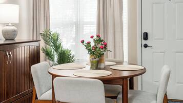 Un comedor con una mesa redonda, cuatro sillas, un jarrón con flores, una lámpara y un aparador.