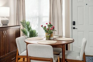 Un comedor con una mesa redonda, cuatro sillas, un jarrón con flores, una lámpara y un aparador.