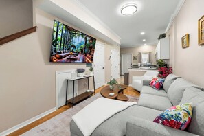 Una sala de estar moderna con un televisor de pantalla plana grande, un conjunto de sofás, una mesa de centro y un comedor con una cocina al fondo.
