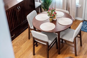 A round wooden dining table with four chairs, a vase with pink flowers, and a wooden cabinet in the background.