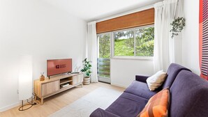 Una sala de estar moderna con televisor de pantalla plana, mueble bajo para televisión de madera, sofá morado y un ventanal con cortinas.