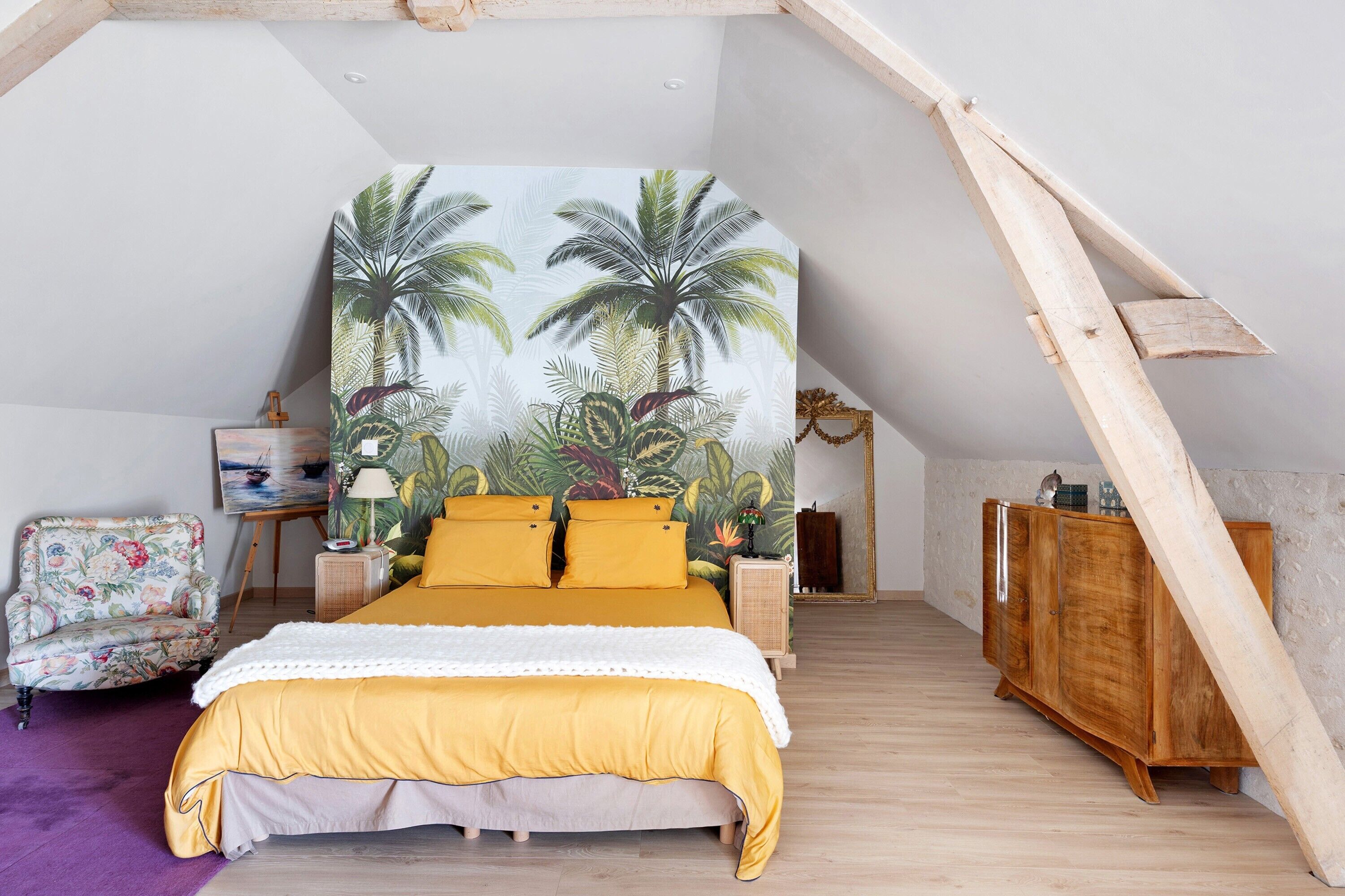 Quarto com teto inclinado, cama com roupa de cama amarela, poltrona floral, armário de madeira e um grande mural tropical na parede.
