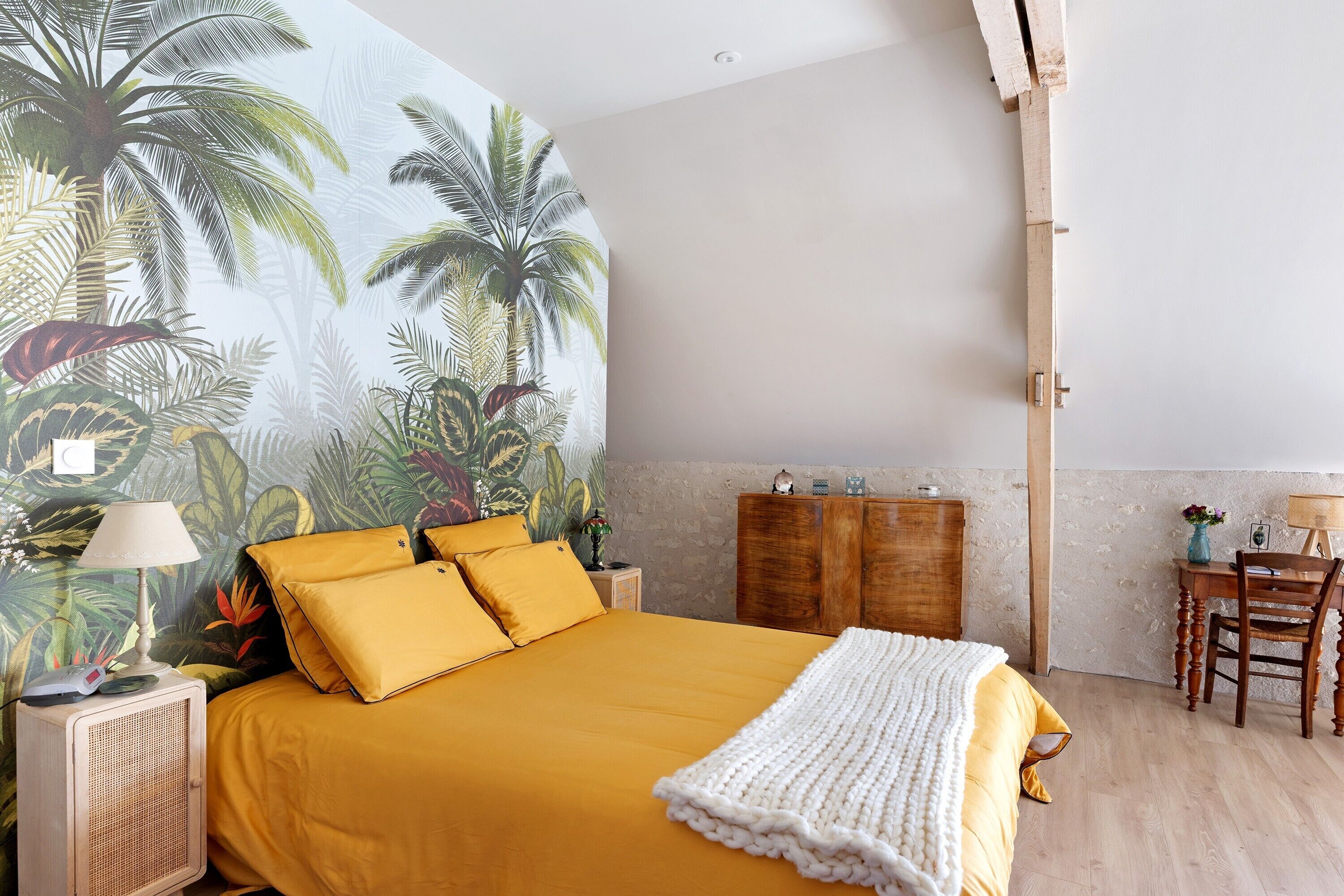 A bedroom with a bed, a nightstand, a lamp, a wooden dresser, and a desk with a chair.