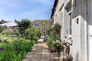 Ein schmaler Steinweg, gesäumt von üppiger Bepflanzung und Blumentöpfen, führt zu einem Gebäude mit weißer Fassade und traditioneller Architektur.