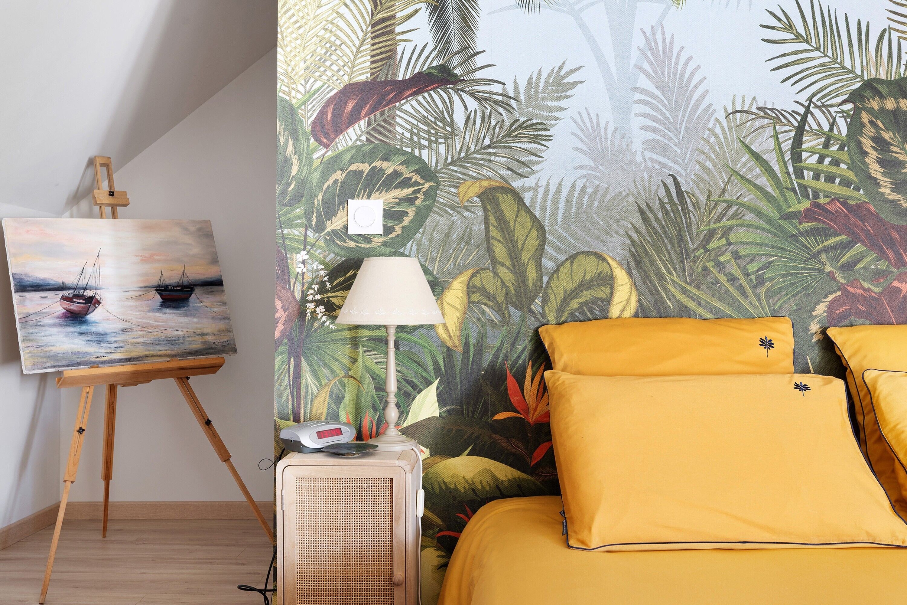 Een kamer met een bed, een schilderij op een staf, een lamp en een muur met tropische behang.