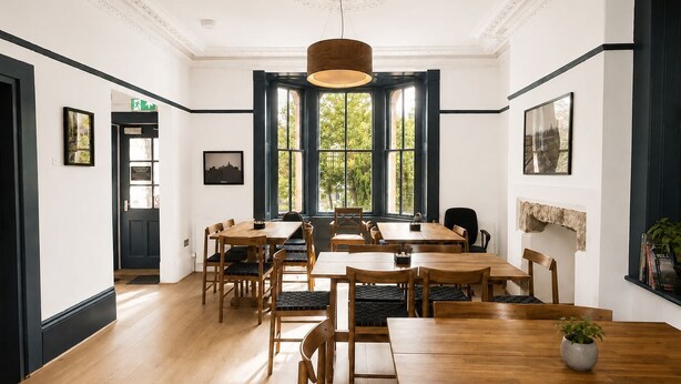 Un'ampia sala da pranzo con tavoli e sedie in legno, grandi finestre vista su spazi verdi e un soffitto decorativo.