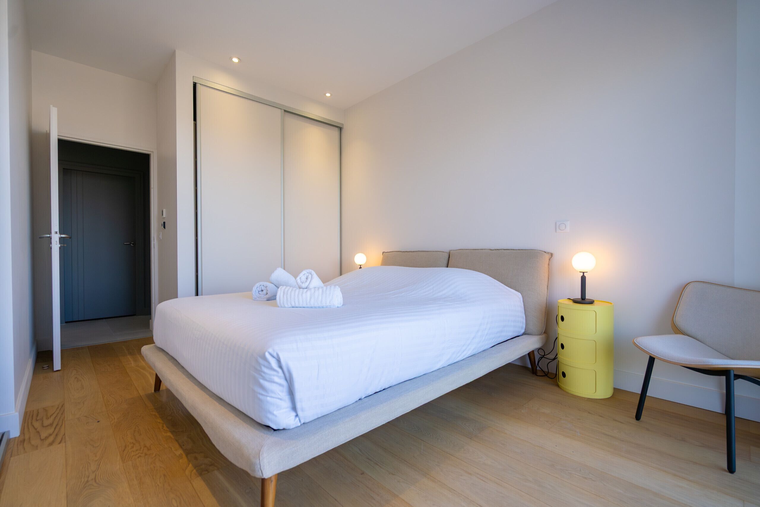 A modern bedroom with a bed, bedside lamp, chair, and a door leading to another room.
