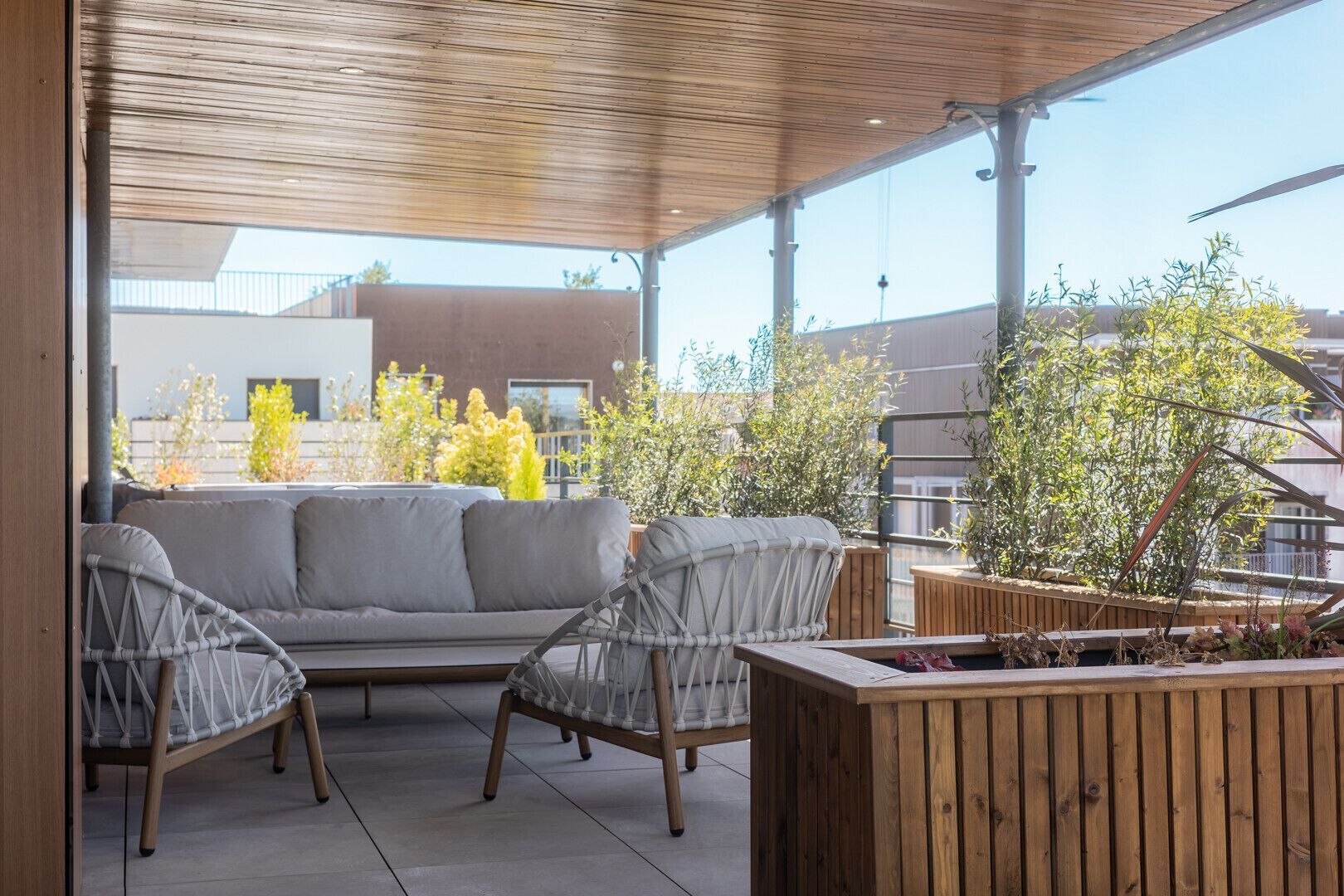Un espacio de estar al aire libre con una mesa de madera y dos sillas, un sofá gris y una jardinera de madera con plantas.