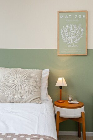 Una cama con un almohadón blanco y un copete, un taburete de madera pequeño con una lámpara, y una obra de arte enmarcada en la pared.