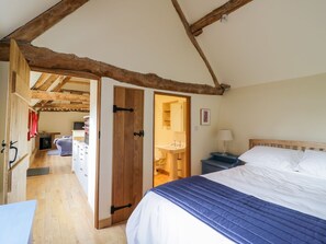 A bedroom with a wooden ceiling, a bed with a blue and white duvet, a bedside table with a lamp, and a bathroom visible through an open door. - Ryepiece (Stratford-upon-Avon)