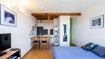 Een slaapkamer met een bed, een televisie, een bureau met stoel, en een kitchenette op de achtergrond.