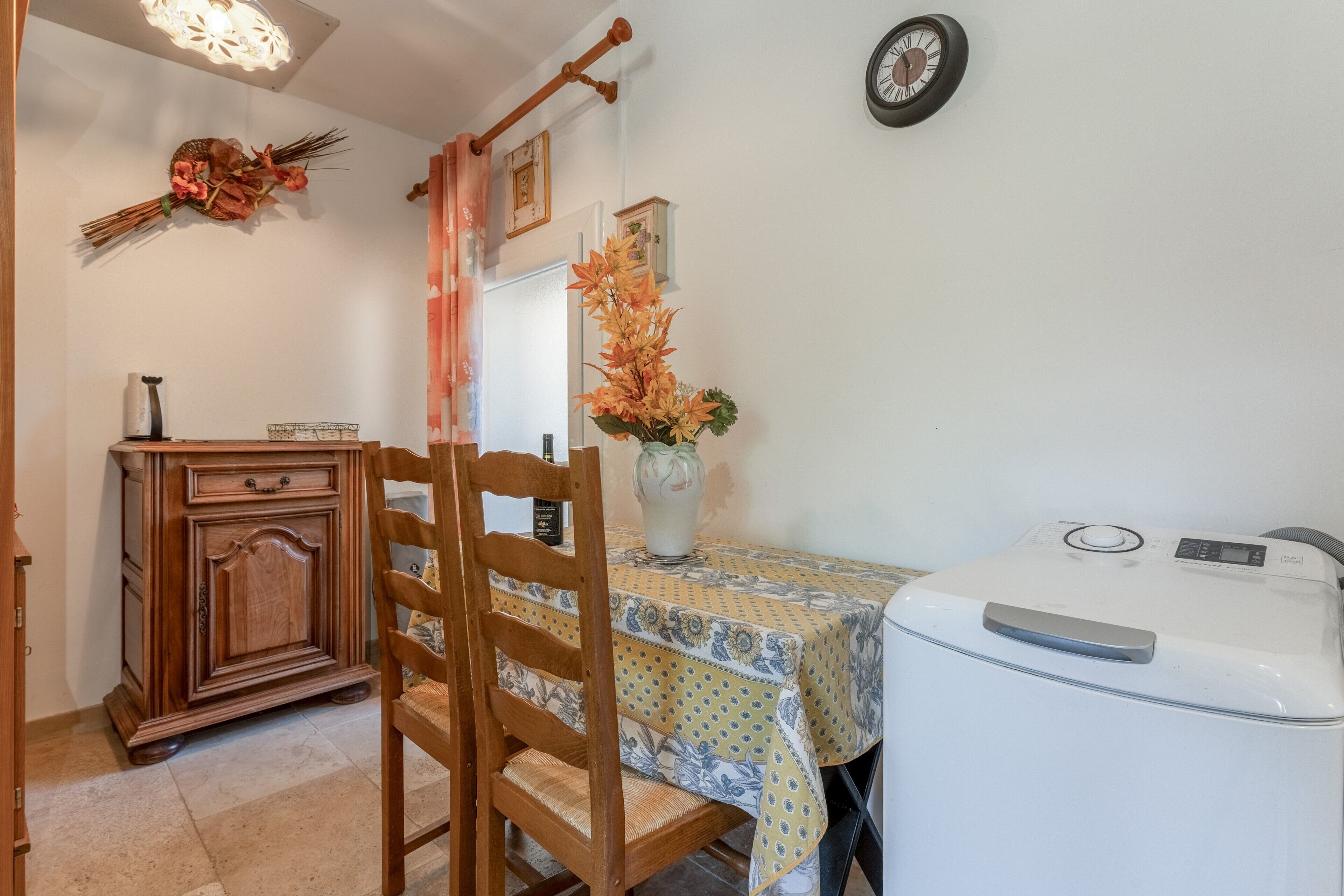 A room with a wooden cabinet, a chair, a table with a patterned tablecloth, a vase with flowers, a clock on the wall, and a white appliance.