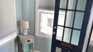 Una habitación con una puerta azul oscuro, una ventana con persianas, una mesita con una lámpara y una alfombra estampada.