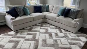 Un sofá seccional color beige con cojines estampados, una alfombra geométrica y ventanas amplias con persianas.
