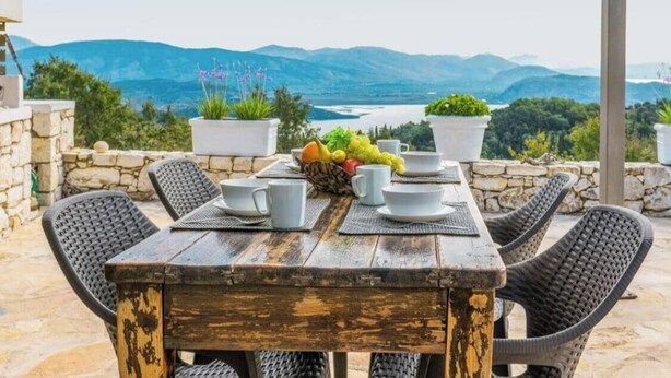Un tavolo di legno con piatti e tazze bianche, circondato da sedie in vimini, su un patio in pietra con piante in vaso e una vista panoramica.