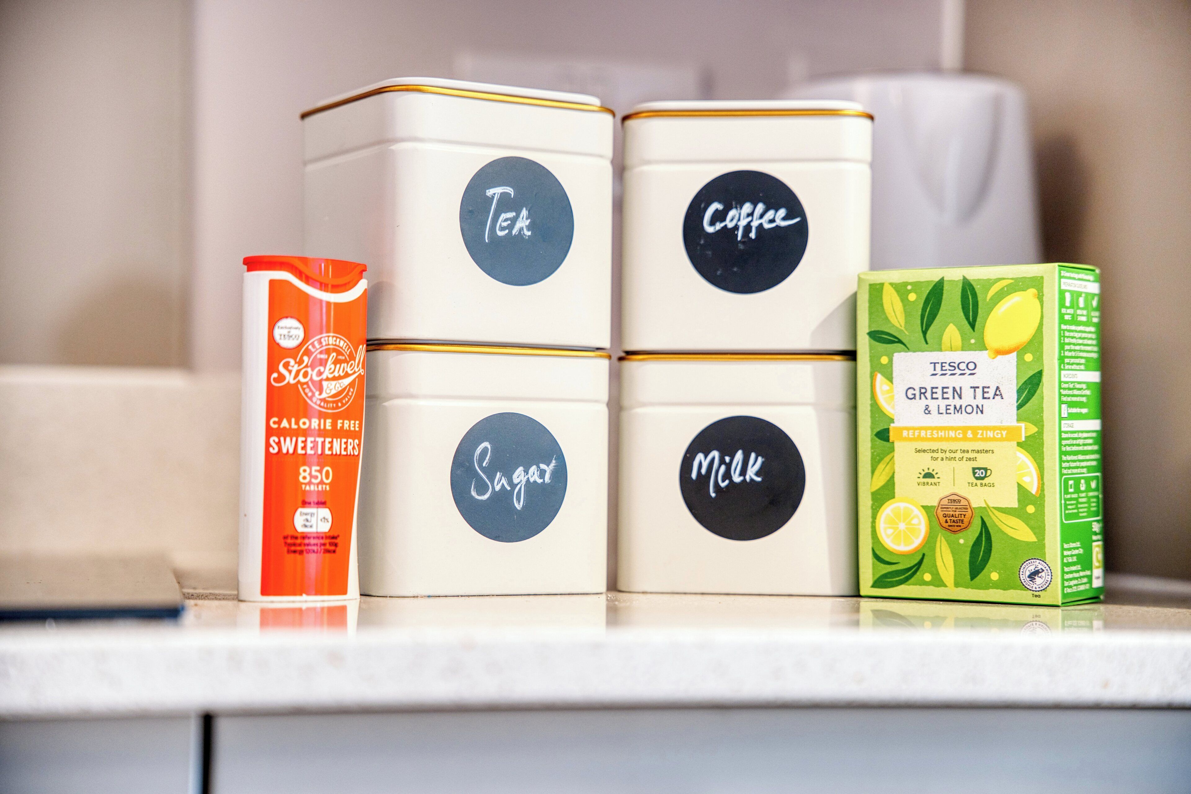 Four containers labeled Tea, Coffee, Sugar, and Milk, and a box of Tesco Green Tea & Lemon.