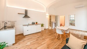 Een moderne keuken met witte kastjes, een inbouwfornuis en een groot eiland. Er is een eethoek met een witte tafel en stoelen, en een woonkamer met een bank en een open haard.