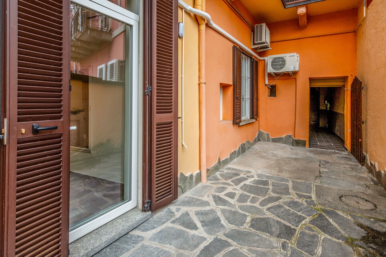 A narrow stone-paved corridor with a glass door and brown shutters, leading to a building with orange walls and air conditioning units.
