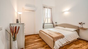 Een slaapkamer met een bed, nachtlampjes, een radiator, een raam met gordijnen, een wandairconditioner en een nachtkastje met een lamp.