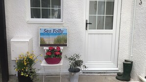 A white door with a 'Sea Folly' sign, a potted plant with yellow flowers, a potted plant with red flowers, a metal stand, and a pair of green boots.