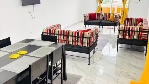 A modern living room with a glass dining table, striped sofas, and a flat-screen TV mounted on the wall. - Résidence au 2 Plateaux 3 Chambres (Abidjan)