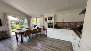 A modern kitchen with white cabinets, a dining area with a wooden table and chairs, and a view of greenery outside. - Chalet in Biddinghuizen, Pool Facilities (Biddinghuizen)