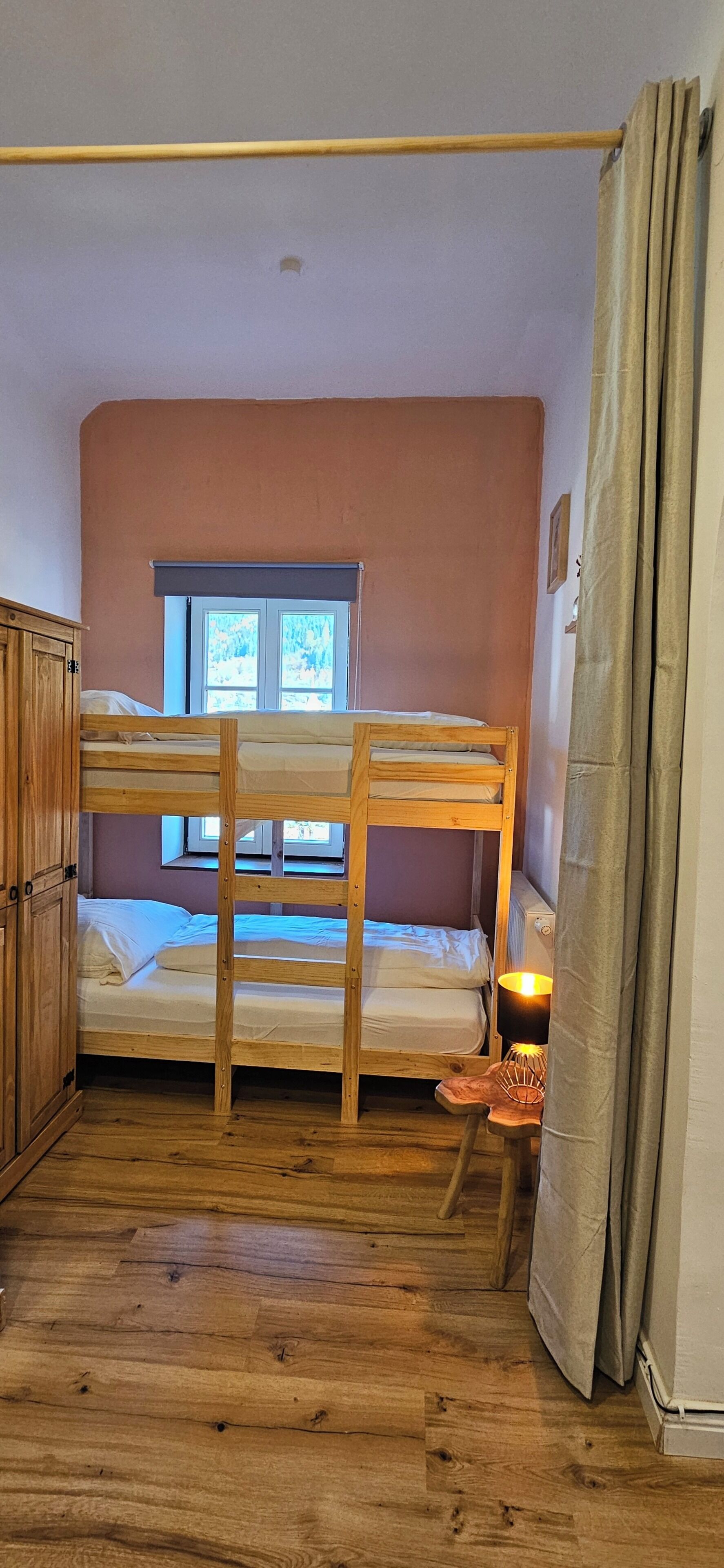 A bunk bed room with a window, wooden floor, and a small table with a lamp.