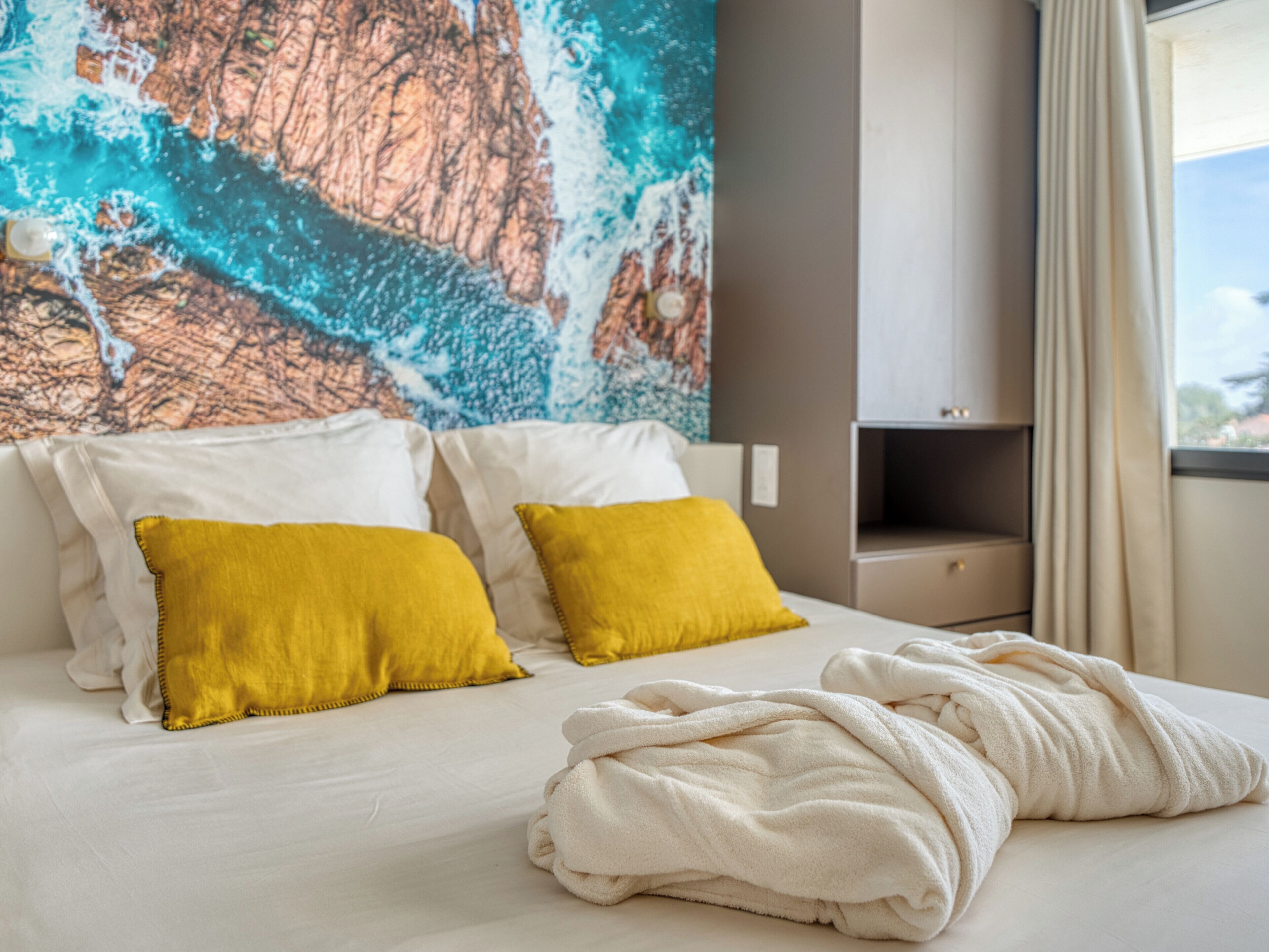 A hotel room with a bed, two pillows, a blanket, a wall with a painting, a nightstand, and a window with curtains.