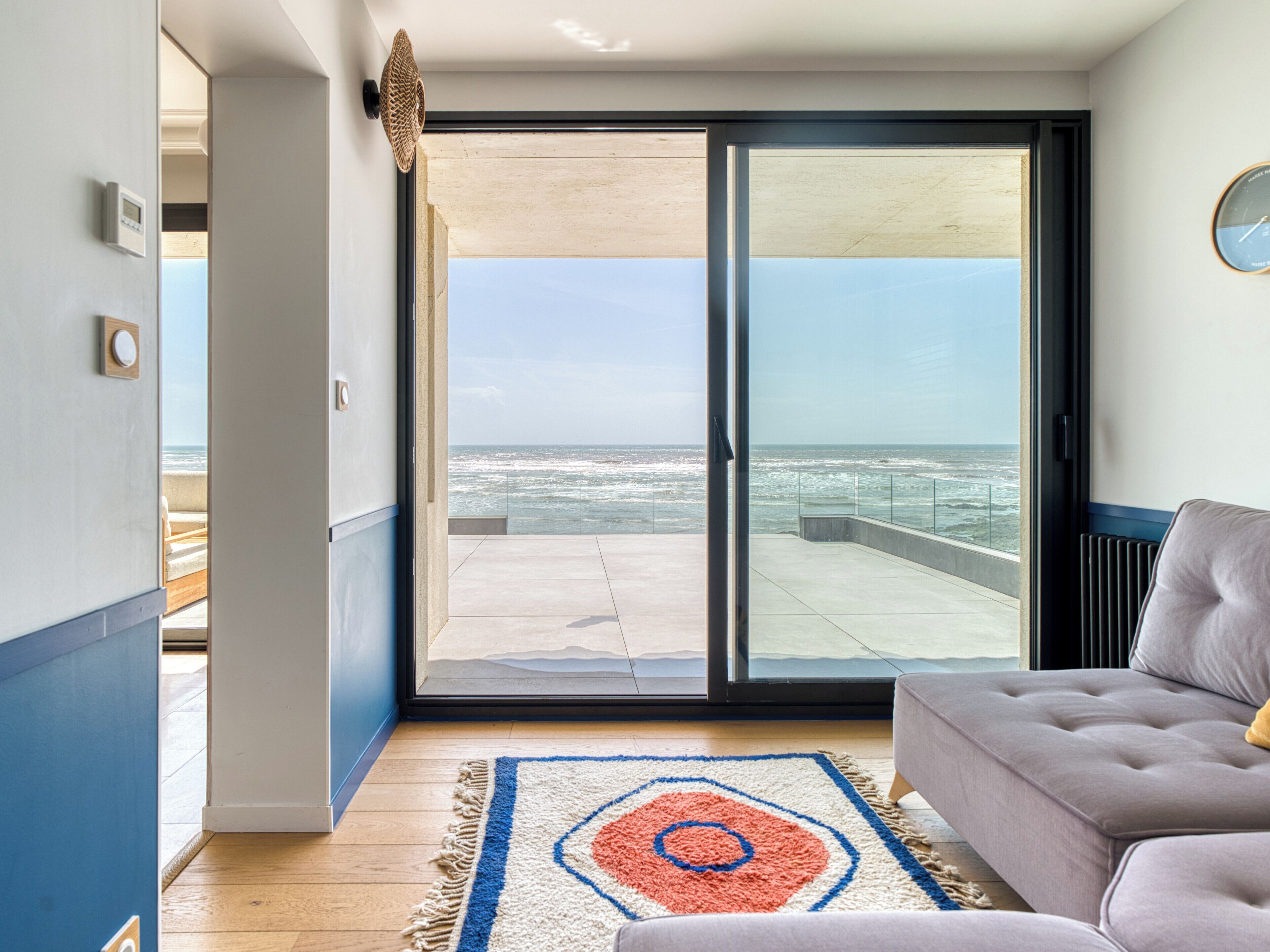 A room with a view of the ocean through a sliding glass door, a sofa, a rug, and a wall-mounted clock.