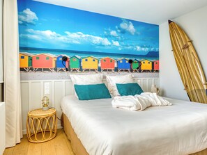 Et soveværelse med en seng, puder, et rundt viklet natbord, en surfbræt og et strandmaleri på væggen.