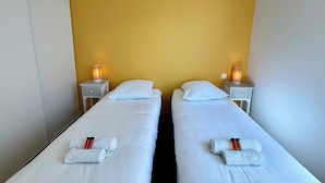 A hotel room with two single beds, each with a white pillow and a rolled towel. There are two bedside tables with lamps and a small drawer. The room has a yellow accent wall and a white ceiling.