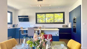 A modern kitchen with blue cabinets, a dining area with yellow chairs, and a large window with a view of trees.