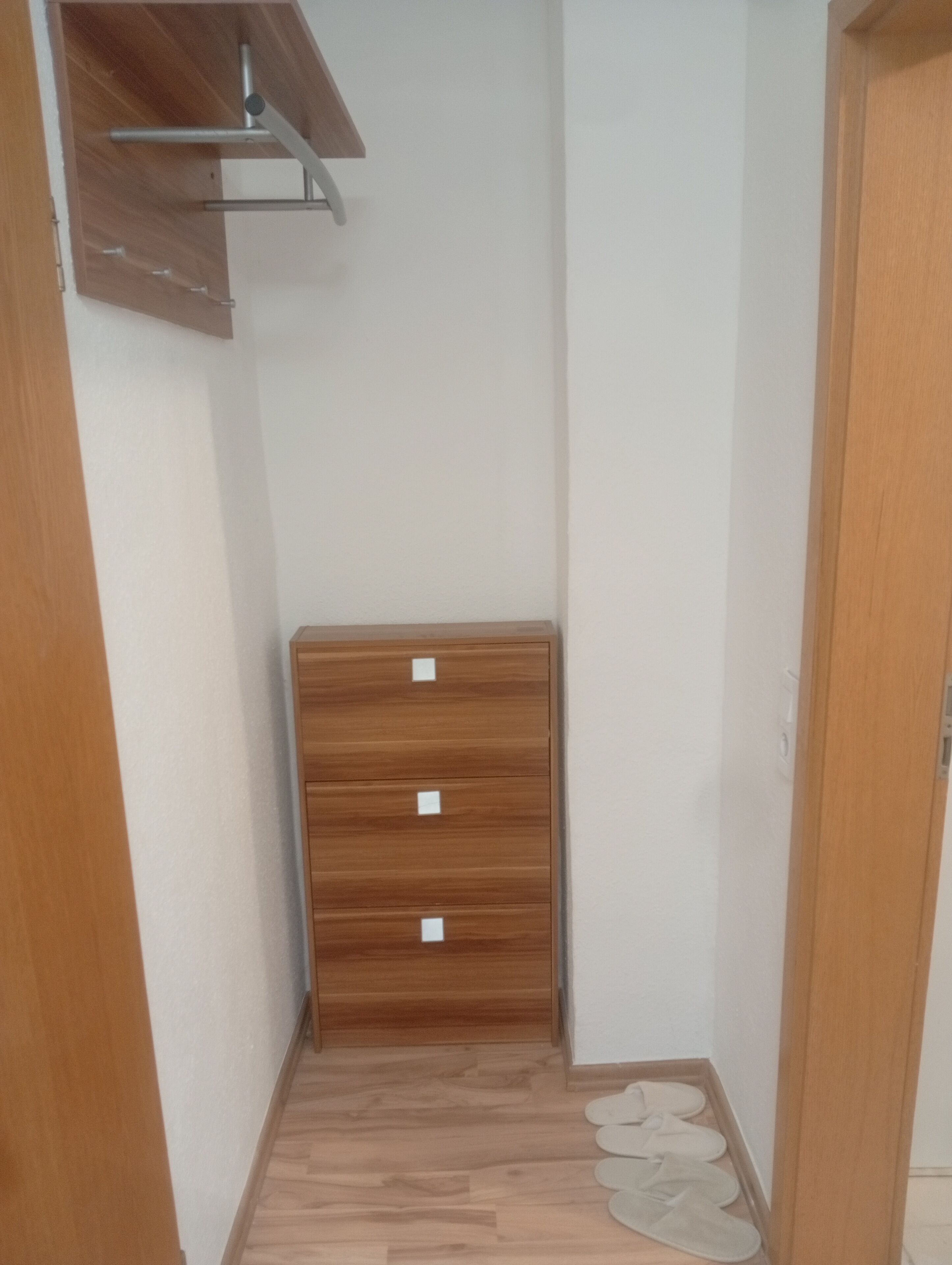 Een smalle gang met een houten garderobe, een metalen plank en een paar slippers op de vloer.